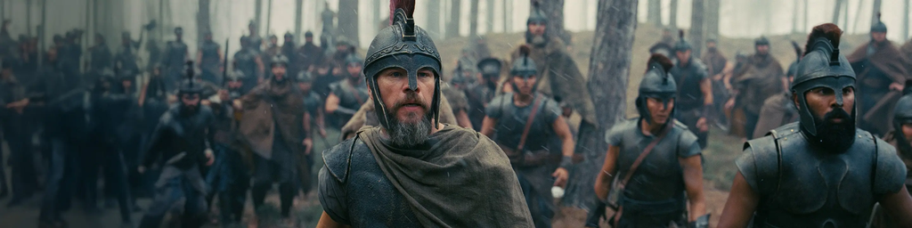Cena do filme A Odisseia, mostrando um grupo de guerreiros medievais avançando por uma floresta. Em primeiro plano, três guerreiros usam capacetes e armaduras, enquanto dezenas de combatentes aparecem ao fundo entre as árvores.